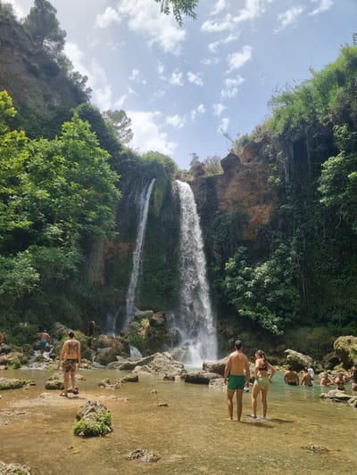In the city of Anna, Spain, a scenic waterfall cascades down a rocky, vegetation-covered cliff face. The water collects in a shallow, clear pool below, where several people are wading and swimming. In the foreground, a man with a large tattoo on his back stands facing away from the camera, wearing patterned swim shorts. To his right, a couple, a man in green swim shorts and a woman in a bikini, stand in the water. Other individuals are visible further in the pool, enjoying the water and the natural surroundings. The landscape is lush with green trees and foliage, contrasting with the rocky cliffs and the bright blue sky dotted with white clouds. The overall scene depicts a popular natural spot for recreation and relaxation.
