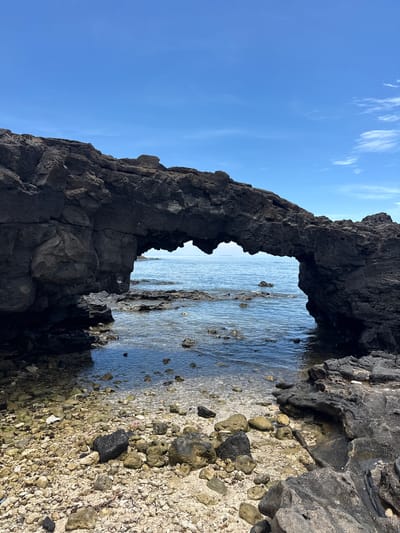 This is a vertical, eye-level, medium shot of a natural rock arch on the coast of Lý Sơn Island, Vietnam. The archway is formed from dark, jagged volcanic rock and frames a view of the calm, blue ocean and a clear sky with sparse white clouds. The foreground is a rocky beach composed of pebbles, sand, and scattered rocks. The water is clear and shallow near the shore, with some smaller rocks visible beneath the surface. The lighting suggests it is a bright, sunny day. There are no people or animals visible in the image.