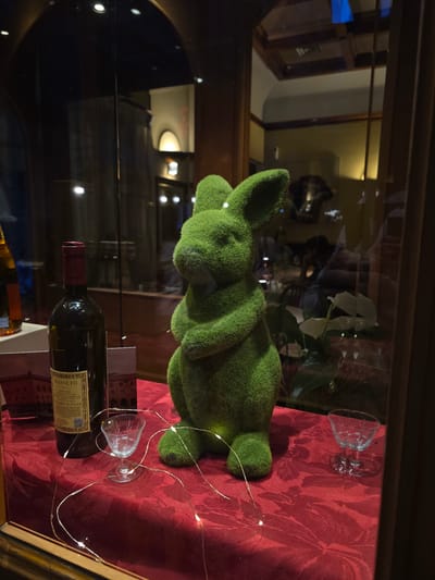 The image displays a close-up view of a window display, likely in a shop or restaurant in Bologna, Italy. In the foreground, a vibrantly green, moss-covered bunny statue stands prominently. To its left is a dark glass wine bottle with a red label, and further left, part of another bottle is visible. Two small, clear glass cups are placed on a red, patterned tablecloth, and thin, luminous string lights are artfully draped around the objects. The background, visible through the glass, suggests an interior setting with wooden paneling and reflections of lights and possibly people seated at tables, hinting at a dimly lit, possibly atmospheric, environment.