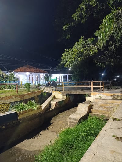 The image shows a concrete drainage ditch at night in Kecamatan Panji, Indonesia (latitude: -7.697, longitude: 114.032). The ditch is lined with concrete and has a bridge over it. The bridge is made of concrete and has a railing. The ditch is partially filled with water. The ditch is surrounded by greenery and concrete. Two figures are sitting on a step near the edge of the ditch. The image was taken at night and is lit by a few streetlights. The building in the distance is lit with bright light. The building in the background is a public building. The building has a tiled roof and windows. The building has a light on at the top and appears to be open for business. There is a large, green bush in the foreground, near the ditch, and there is a white light pole in the background. There are wires strung across the image in the background. The image was taken at night.