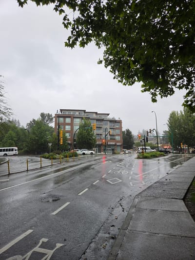 The image depicts a rainy day scene on a street in North Vancouver, Canada. The foreground is dominated by a wet asphalt road, marked with white lines indicating a bike lane and traffic lanes. A sidewalk made of concrete slabs runs parallel to the road, also appearing wet. Yellow bollards line the edge of the road.
In the mid-ground, a modern brick and glass apartment building stands tall, with balconies and numerous windows. Across the street, trees line the sidewalk, their leaves a vibrant green. Traffic lights are visible, some showing red, indicating a halt in traffic. A white bus is parked on the side of the road.
The background shows more trees and buildings, with a misty or cloudy sky suggesting overcast weather. The overall mood is subdued due to the rain and the muted colors of the cloudy sky. The presence of the bike lane and the bus suggest an urban or suburban setting. No people are clearly visible in the image.
A street sign is visible on the apartment building, reading "Marine Drive". The time of day appears to be daytime, but the heavy cloud cover makes it difficult to determine the exact time. The wet conditions are the most prominent feature, suggesting recent or ongoing rainfall.
