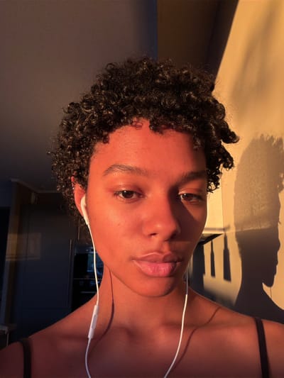 A young, dark-skinned individual with curly hair is facing the camera with earphones in. They are in a dimly lit room, likely a kitchen, with a grey wall to their left and a counter and cabinets visible in the background. The right side of their face is bathed in warm light, creating a strong contrast. The shadows suggest there might be a window to the right with a patterned blind, casting shadows on the wall and counter. The image is tilted to the side. There is no indication of the location being Champs-sur-Marne, France.