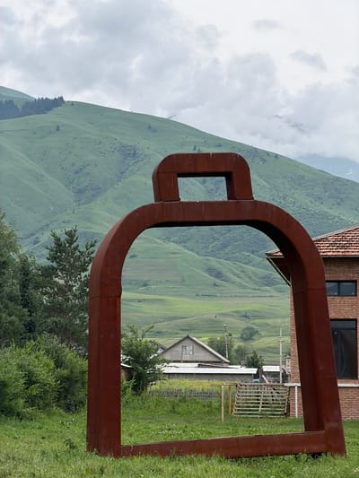 The image shows a large, rusty, abstract sculpture in the foreground. The sculpture is U-shaped and made of thick, square metal beams. Through the sculpture, a rural landscape of rolling green hills is visible. There are a few houses and other buildings nestled in the hills. In the far distance, a cloudy sky looms over the mountains. The time of day appears to be daytime, likely overcast given the cloud cover. The overall setting is a serene, mountainous countryside. There is no visible text in the image.