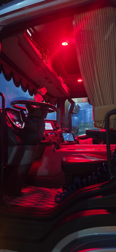 The image shows the interior of a truck cabin, illuminated by red ambient lighting. The driver's seat and dashboard are visible, with a steering wheel, control panels, and screens. Two dice hang from the ceiling near the windshield. A curtain is drawn to the right, and the floor has a patterned mat. The scene is consistent with the interior of a truck parked in a garage or workshop, potentially in Épila, Spain, given the request, though no explicit urban features are visible within the cabin.