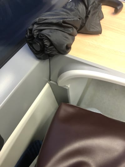 An umbrella, collapsed and black, sits on a light wood surface next to a curved white partition. A brown leather bag hangs over a lower level, obscuring details beneath. The scene's surfaces are light gray, with hints of navy blue below the umbrella. The lighting is bright, emphasizing the texture and color variations across the items. The composition is tightly framed, focusing on the juxtaposition of these everyday objects.