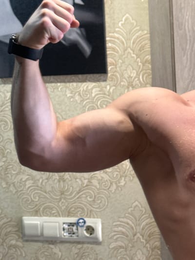 A close-up, slightly rotated image shows a muscular arm flexed. The background features patterned wallpaper and a white electrical outlet with switches. A black wristband is visible on the wrist of the flexed arm. A partial view of a black and white photo can be seen on the left. The location is likely indoors, but there is no discernible evidence in the image to place it in Dobryanka, Russia.