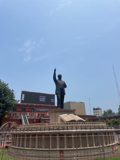 The image shows a statue of B.R. Ambedkar, a prominent figure in Indian history, located in Amritsar, India. The statue stands on a pedestal which includes an open book, symbolizing knowledge and education. Behind the statue, a large brick building houses several businesses, including "Barista" and "The Coffee Bean & Tea Leaf," as well as a Starbucks. A large digital billboard is also visible on the building, displaying numbers and text. The scene is set against a clear blue sky with a few wispy clouds. In the background, several tall buildings and communication towers can be seen, indicating an urban environment. The foreground features a metal railing and lush green grass. The overall impression is that of a public monument in a bustling city.