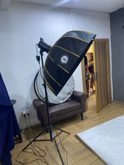 This media file presents an indoor scene, rotated 90 degrees to the left, appearing to be a temporary photography setup in Animashawun, Nigeria.
The primary subjects are photography equipment arranged in a room. In the foreground, dominating the left side of the frame, is a large black photography softbox with prominent gold stripes and a white circular "DC" logo. Attached to it is a strobe light, partially visible, with "SK300" and likely "GODOX" text on its body. Below and slightly behind the softbox, a circular silver photography reflector stands on the floor.
To the right of the softbox and reflector, a brown leather two-seater sofa is visible. Extending from the bottom right towards the center of the image is a black light stand or boom arm, with various power cords trailing from it to an electrical outlet mounted on the light-colored wall.
In the background, there's a wooden structure, possibly a door or a large cabinet, positioned above the sofa. Behind this wooden element, a pile of patterned textiles or clothing (brown and floral) is discernible. The floor is covered with light-colored wooden planks, and the walls are white or off-white, with a darker ceiling visible in the upper left corner. The setup suggests that a photography session is either being prepared, is in progress, or has recently concluded in this space. No people are visible in the image.