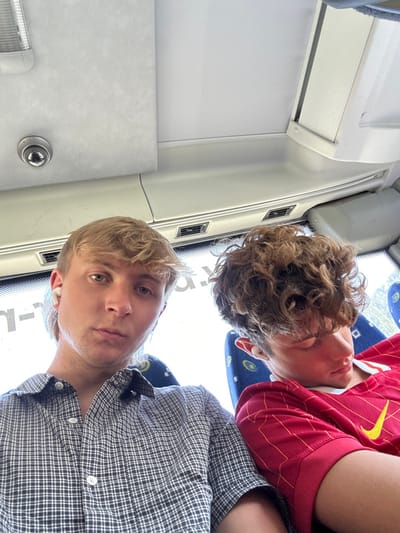Two young men pose for an upward-angled selfie inside a vehicle, likely a bus. The man in the foreground wears a plaid shirt and a neutral expression. The other man, resting behind him, has curly hair and wears a red and yellow jersey. The interior shows white walls, air vents, and patterned blue seats, suggesting a public transportation setting.