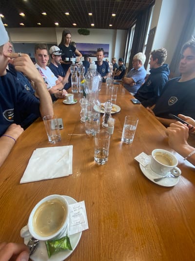 A group of approximately a dozen people are seated along a long, wooden table. Many are wearing similar navy blue athletic wear. Empty water bottles and water glasses litter the center of the table. Some individuals are drinking coffee in white cups. Sugar packets and paper napkins are on the table. The image suggests a meeting, gathering, or break taking place at a cafe, possibly after a sporting event, in Bělá pod Pradědem, Czechia.