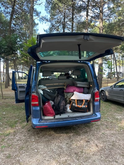 The image shows the open trunk of a blue Volkswagen Transporter, heavily packed with luggage and camping gear. An "Icepeak" branded bag is visible among the items. The rear passenger-side sliding door is also open. The car is parked in a wooded area with a gravelly surface, likely a campground. A silver car is parked behind the blue van. The location is Donji Štoj, Montenegro, indicated by the GPS coordinates. The environment appears to be sunny, with tall pine trees visible in the background. No people are visible in the image.