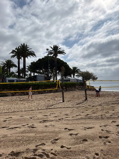 This image depicts a beach volleyball court on a sandy beach in Mont-roig del Camp, Spain, under an overcast sky. Two male individuals are present on the court. On the left, a fair-skinned male, shirtless and wearing white shorts, stands barefoot on the sand, facing towards the right side of the court in an active stance. On the right, a second fair-skinned male, wearing a dark shirt and shorts, crouches near the net, holding a red and white volleyball, poised to serve.
The volleyball net, bearing "COPAYA" branding on its yellow tape borders, is stretched between two rough-hewn wooden poles. The sand in the foreground and on the court shows numerous footprints and textured patterns, possibly from beach grooming. In the background, the Mediterranean Sea is visible, appearing calm with a subtle horizon line.
Behind the beach, a low, rocky embankment topped with dense green foliage and smaller, gnarled trees separates the sand from an area containing several parked recreational vehicles (RVs) or caravans. Notable vehicles include a white "KNAUS" branded RV and a darker "COPAYA" branded RV, with other vehicles partially obscured by tall palm trees that punctuate the skyline. A streetlamp is also visible above the foliage. The sky is largely covered by grey stratocumulus clouds, with some lighter patches indicating thinner cloud cover or breaks. Two additional human figures are visible as dark silhouettes on the wet sand near the water's edge in the far background, one standing and one further to the right.