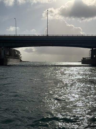 This photograph depicts a view under a bridge in Bal Harbour Village, United States of America. The left side of the frame showcases the structure of the bridge, with its support beams and a couple of lampposts visible against a cloudy sky. The right side is dominated by the rippling surface of the water, reflecting sunlight and creating a glittering effect. In the distance, there's a glimpse of land with some vegetation and a tall structure, possibly a building. The overall tone of the image is muted, with a predominance of grayscale tones.