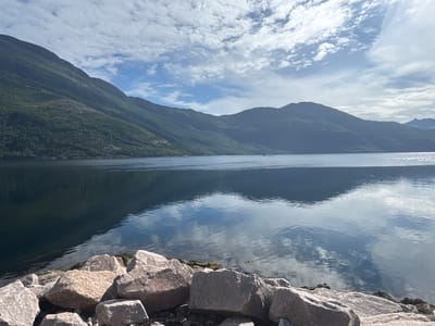 This image shows a serene natural landscape with a large body of water in the foreground, flanked by mountains. The water is calm and reflects the sky, which is partly cloudy with patches of blue. The mountains are covered in lush green vegetation, suggesting a temperate climate. Large rocks are scattered along the shoreline in the foreground, adding texture to the scene. The overall impression is one of tranquility and natural beauty, consistent with the scenery found in Sund, Norway, known for its dramatic fjords and mountainous terrain. There are no people or man-made structures visible in the image, emphasizing the wild and untouched nature of the environment. The lighting suggests it is daytime, possibly late afternoon, given the soft quality of the light and the long reflections on the water.