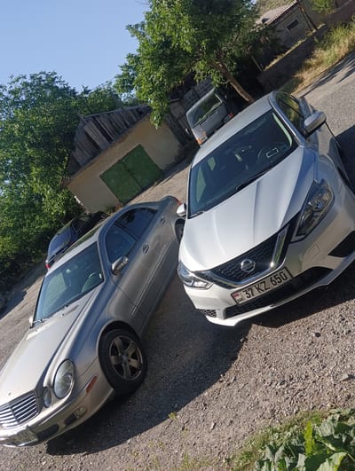 In Talin, Armenia, on a gravel lot under a clear blue sky, two sedans are parked. The vehicle in the foreground is a silver Nissan Sentra with license plate "37 KZ 650". To its left and slightly behind is a silver Mercedes-Benz E-Class with license plate "67 CV 599". In the background, a weathered wooden building with green doors sits adjacent to a dirt road and lush green trees. A dark-colored van is also visible parked behind the Mercedes-Benz. The scene is illuminated by bright sunlight, casting distinct shadows.
