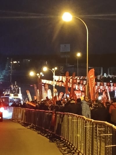The image shows a crowd of people standing behind a metal barrier on a street in Istanbul, Türkiye at night. The crowd is facing a row of large red and white flags strung across the street. The street is empty in front of the crowd. There are street lights, some with multiple lights, illuminating the scene. There are also vehicles in the distance. The scene is located at the coordinates 41.084, 28.678 in Istanbul, Türkiye.