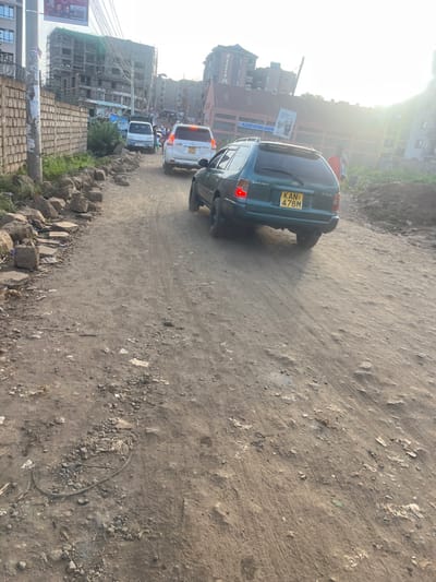 The image depicts an unpaved, dusty dirt road in Gitothua ward, Kenya, illuminated by bright daylight, with strong sunlight emanating from the upper right. The road surface is uneven, showing ruts, loose gravel, and scattered debris.
In the mid-ground, a dark green Toyota Corolla station wagon (license plate KAN 478M) is angled away from the camera, positioned on the left side of the road, appearing to be in motion or recently stopped, with its brake lights illuminated. Behind it, a white SUV, possibly a Toyota Land Cruiser Prado, is visible, followed by a white van further in the distance. These vehicles are aligned, suggesting traffic flow.
On the left side of the road, a low wall constructed from large, irregular stones lines the edge. Behind this wall, a wooden utility pole with multiple overhead wires stands. Further back, multi-story buildings are visible, some appearing to be under construction with scaffolding and green netting, while others are completed structures. A large banner with "BLESSED NATIONS ASSEMBLY" is affixed to one building.
On the right side of the road, a dirt embankment rises, characterized by loose soil and sparse dry vegetation. A person, indistinct due to distance and lighting, is standing or walking near this embankment, wearing a reddish-orange upper garment. The foreground consists entirely of the uneven, dusty road surface with minor debris.