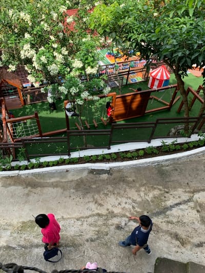 The scene shows a fenced-in playground area in Kecamatan Borobudur, Indonesia. The playground is made of artificial grass with a wooden structure in the background. There is a red and white striped tent in the upper-right corner of the playground. A girl in a red shirt is standing next to a wooden structure in the middle of the playground. Three people are standing on a concrete pathway outside of the playground. A woman in a pink shirt is looking down at the ground and a man in a blue shirt is looking down at the ground. There is a child, obscured by the pathway's edge, wearing a pink and white shirt in the bottom-center of the image. The playground is surrounded by a white concrete curb and a black metal fence, along with a row of green shrubs. There is a large green tree with white flowers in the upper-left corner of the image. This scene is located at a latitude of -7.606 and a longitude of 110.18.