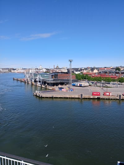 This is an image of the West Harbour terminal in Helsinki, Finland, on a clear day with a bright blue sky and a few wispy clouds. The foreground shows the dark blue, slightly rippled water of the Baltic Sea, with a white railing in the bottom left corner. Several seagulls are visible in the water and in the air. The middle ground features a modern ferry terminal building with a white, multi-story structure and large glass windows. Adjacent to the terminal are several docked ferries or parts of ferries, along with cargo containers, including one red container labeled "VIKING LINE." A tall metal tower with lights stands near the terminal. In the background, a city skyline is visible, characterized by various buildings of different heights and architectural styles, including domes and spires, suggesting a mix of historical and contemporary architecture. Trees are also present along the waterfront and behind the terminal. The ground area around the terminal appears to be a paved parking lot with some vehicles.