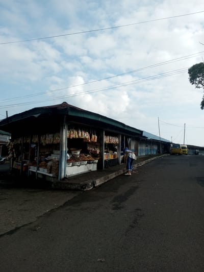 The image depicts a street scene in Mya Sit Kaing Ywar Thit, Myanmar. A row of small, covered market stalls lines the left side of the asphalt road. The first stall is fully stocked with various snacks and food items, many of which are packaged and hanging. A person, possibly a customer, is standing in front of this stall, looking at the goods. Farther down the road are additional stalls, with a yellow vehicle parked in front. A person on a motorcycle is also visible in the distance. The sky is partly cloudy, with power lines running overhead.