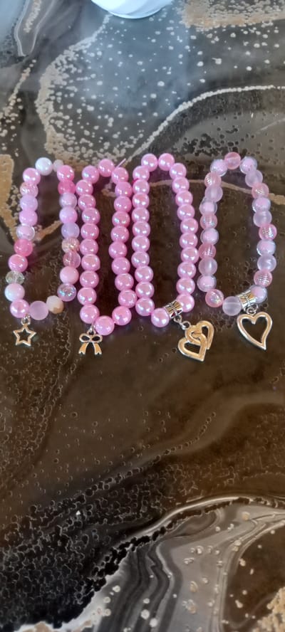 Four pink beaded bracelets are laid out on a dark, glossy surface. The beads vary in size and opacity, with some appearing iridescent and others frosted. Each bracelet is adorned with a silver-toned charm: a star, a bow, two intertwined hearts, and a single heart. The surface has a marbled or abstract pattern with lighter and darker areas and small, raised circular textures. The lighting is soft, casting subtle shadows. The image does not depict any recognizable city landmarks or elements that would allow for specific location identification within Iskra, Bulgaria.