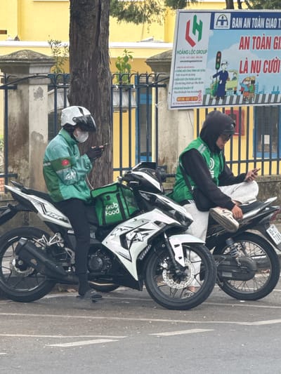 Two food delivery drivers on motorcycles are parked on a paved street, likely in Xuân Hương - Đà Lạt, Vietnam, as suggested by the billboard in the background. The driver on the left is wearing a turquoise jacket with a Grab Food logo and a white helmet. They are looking down at their phone. The motorcycle is white and black. The driver on the right is wearing a black hoodie with a green vest and a black helmet, also looking at their phone. Their motorcycle is black. In the background, a billboard with Vietnamese text and graphics about traffic safety is visible. The environment appears to be outdoors, with a building and a tree visible behind the drivers.