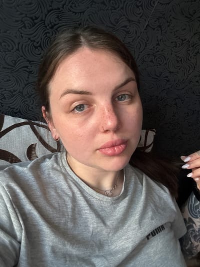 A young woman is captured in a close-up, top-down selfie while lying down in an indoor setting in Chornomorskoe, Ukraine. She has long dark brown hair, blue-grey eyes, and a small hoop nose piercing. Her lips are a light pink, and freckles are visible on her nose and cheeks. She wears a light grey t-shirt with a visible, reversed "PUMA" logo across the chest, suggesting a front-facing camera or mirror shot. A delicate silver chain necklace adorns her neck. Her fingernails are long, almond-shaped, and adorned with a light, decorative pattern, possibly white marble or glitter. A dark, intricate tattoo sleeve is visible on her right arm/shoulder area. She gazes directly at the camera with a neutral expression. The background consists of a dark grey or black patterned fabric, likely a bedspread or sofa, featuring swirling, ornate designs. Another section of fabric with a lighter grey background and brown geometric patterns is also partially visible, possibly part of a cushion. The overall impression is one of a relaxed, informal moment.