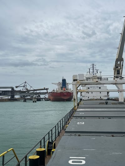 This forensic image captures a maritime industrial scene in Uribia, Colombia, under a uniformly overcast sky. The viewpoint is from the deck of a large cargo vessel, looking forward and towards the port side. The foreground displays the gray, textured deck with several large hatch covers featuring prominent white numerical markings, specifically "10" and "20". A dark metal railing with a yellow top runs along the left edge of the deck, terminating near black cylindrical bollards topped with yellow caps. The water adjacent to the vessel is a murky teal color.
In the midground, to the right, a white cargo ship, identified by the name "HELD SHIPPING" on its superstructure, is visible. This vessel features a complex array of antennas and radar equipment on its mast and a white crane arm marked with "SWL 20.5T". Towards the center-left midground, a large red-hulled bulk carrier, displaying "NAVIOS RAY" and "PANAMA" on its stern, is positioned in the water, appearing to be either anchored or navigating. Adjacent to this vessel on the far left, a substantial industrial bulk loading facility extends over the water, characterized by multiple interconnected metal conveyor structures supported by concrete pylons. No human figures are discernible within the frame.