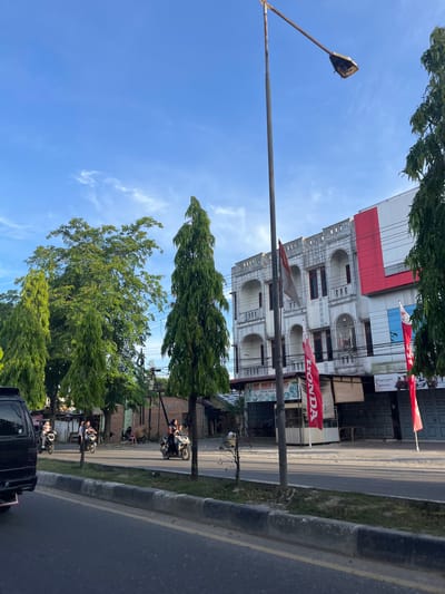 The image captures a street scene in Langsa, Indonesia, under a clear blue sky with scattered wispy clouds. The foreground shows a paved road with a gray concrete barrier and a grassy median. Several trees line the roadside, including tall, slender coniferous trees and a broader deciduous tree.

On the right side of the street, a multi-story building with a mix of architectural styles is visible. The upper floors feature a pale, ornate facade with arched openings and decorative elements, while the lower floors are modern with red and white panels. Red banners with white lettering, one clearly displaying "HONDA," are prominently displayed in front of the building. An Indonesian flag is also seen attached to a pole.

Several people are riding motorcycles on the road. On the left, a black van is partially visible, and two motorcycles with riders are passing. Further down the road, another motorcycle with two passengers is in motion. In the background, more buildings and trees are present, suggesting a commercial or residential area. A street lamp with a broken light fixture stands tall near the center of the image. The lighting indicates it's likely late afternoon or early morning, with the sun casting long shadows.
