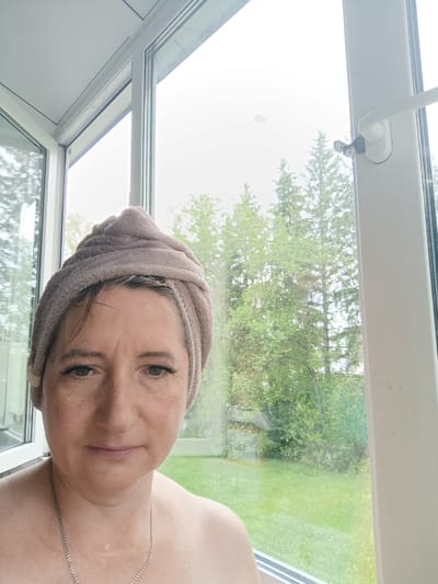 A medium shot captures a woman with a towel wrapped around her head. She has fair skin, light brown hair peeking out from the towel, and green eyes. She is wearing a silver necklace. Her expression is neutral, and she is looking directly at the camera. She appears to be indoors, possibly in a sunroom or on a balcony, with a large window behind her offering a view of green trees and grass. The window frame and a part of a window latch are visible on the right side of the image. The natural lighting suggests it is daytime. The scene appears to be located in Tugulym, Russia.