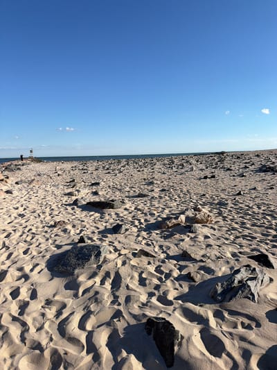 The image depicts a wide, sandy beach with numerous rocks scattered across the surface. The sand is textured with footprints and wave-like patterns. In the background, the ocean stretches to the horizon under a clear blue sky with a few wispy clouds. On the left side of the image, a rocky pier extends into the water, with two individuals standing near a navigational marker. The scene is bathed in sunlight, casting long shadows across the sand. The location is identified as a beach in Hampton Bays, United States of America.