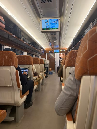 The image depicts the interior of a modern train carriage, likely in Dodoma, Tanzania, given the context of the Tanzanian train network. The carriage is observed from a low angle, looking down the aisle between rows of seats. The seats are upholstered in a cream color with an orange geometric pattern. Luggage is stored on overhead racks. A screen mounted above the aisle displays information, with text visible including "DEST: DAR ES SALAAM." Several passengers are seated, with one individual in the foreground on the left using a tablet. Other passengers are visible further down the aisle, some appearing to be looking forward or engaging with their devices. The overall environment appears clean and well-maintained, suggesting a relatively new or recently refurbished train.