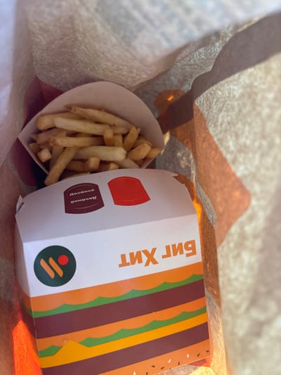 The image shows a fast-food bag with french fries and a burger box inside. The burger box has a depiction of a burger and text that reads "Биг Хит" (Big Hit) in Russian. There is also a circular logo with orange and red elements on a dark green background. A smaller circular label on the box contains Russian text reading "Двойной чизбургер" (Double Cheeseburger). The scene appears to be outdoors, with natural light casting shadows and illuminating the paper bag. The location is identified as Fedorino, Russia.