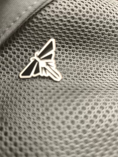 The image shows a silver pin in the shape of a dragonfly, pinned to a dark grey mesh fabric. The pin is located at the upper right corner of the image. The mesh fabric has a repeated pattern of small, hexagonal holes. The coordinates (latitude: 37.982, longitude: 23.656) indicate a location in Koridallos, Greece, but the image does not provide any information about the specific location within the city. The image is taken at a close-up angle, focusing only on the pin and the surrounding fabric. No people, objects, or actions are visible in the image.
