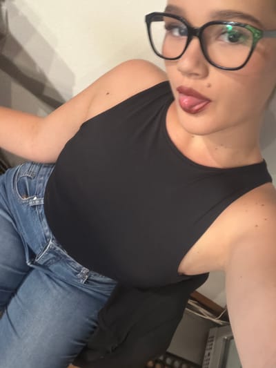 The media file shows a medium shot of a person from the chest up, angled away from the camera. The person is wearing a black tank top and blue jeans. They have on black-rimmed glasses and their tongue is sticking out slightly, with their lips appearing to be glossed. The background is partially visible and shows a wall and what appears to be a printer or scanner on a shelf. There are no visible text elements in the image. The lighting suggests an indoor setting, possibly during the day or evening.