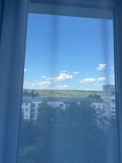 The image shows a view through a window, likely from an apartment building in Strășeni, Moldova. The orientation of the image suggests it was taken vertically. The scene depicts a bright, clear day with a vibrant blue sky dotted with fluffy white clouds. In the middle ground, there is a lush green landscape, possibly fields or a park, stretching towards the horizon. On the right side of the image, there are multi-story buildings, characteristic of an urban or suburban environment. Trees with green foliage are visible, partially obscuring some of the buildings. The buildings appear to be residential or office blocks, with visible windows. The overall impression is one of a pleasant, sunny day. There is no visible text in the image, nor are there any people or specific activities depicted. The light suggests it is daytime, likely mid-morning or afternoon, given the brightness and the angle of the sun. The view out of the window implies the photographer is indoors, looking out.