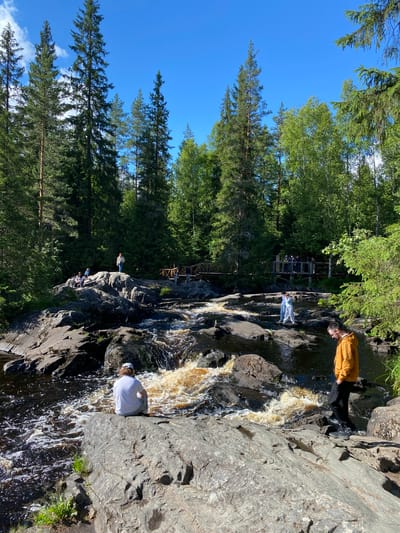 A wide, rocky waterfall flows through a dense forest with tall evergreen trees under a clear blue sky. Several people are present in the scene, interacting with the environment. On a large, flat rock in the foreground, a person wearing a white t-shirt and a white cap sits facing away from the camera, looking towards the turbulent water. To the right of this rock, another person wearing a yellow hooded jacket and black pants stands, looking down at the rocky terrain. In the mid-ground, a pair of people in white outfits stand on rocks in the middle of the stream, appearing to be posing for a photo or observing the water. Further back, on a higher rocky outcrop, a small group of people are gathered, with one person standing near a wooden bridge that spans across the waterfall in the background. More people are visible on the bridge. The environment is natural and undeveloped, with rough rocks and lush green foliage surrounding the cascading water. This scene is characteristic of the Ruskeala Mountain Park in Ruskeala, Russia, known for its marble canyon and natural waterfalls.
