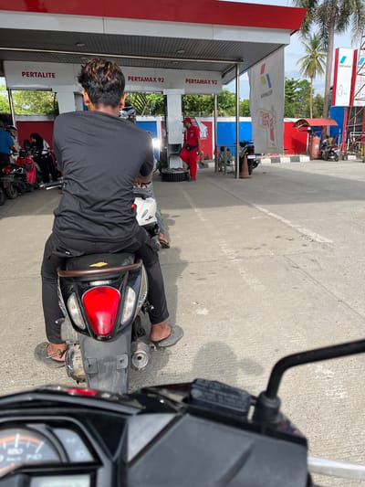 The image depicts a gas station in the city of Barat Yaman, Indonesia. In the foreground, a person is seated on a motorcycle, facing away from the camera. The motorcycle is black with a red taillight and the word "HONDA" visible on its rear. The person is wearing a black t-shirt and black pants, with grey flip-flops. The motorcycle is positioned on a concrete surface, with a white line marking a lane.

In the background, the gas station's canopy is visible, with signs indicating "PERTALITE" and "PERTAMAX 92." A person in a red uniform, likely a gas station attendant, is standing near a fuel pump. Several other motorcycles are parked in the background, some with riders. The environment appears to be outdoors with palm trees visible in the distance, suggesting a tropical climate. The overall scene is a snapshot of everyday life at a gas station in Indonesia.