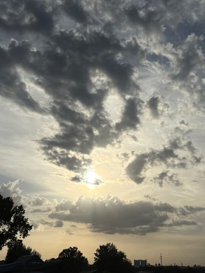 The image depicts a sky filled with clouds, with the sun visible through a gap in the cloud cover. The lower right portion of the image shows silhouettes of trees and rooftops. In the far distance along the horizon, utility poles and buildings are faintly visible. The sky transitions from lighter, brighter areas around the sun to darker, more opaque cloud formations. The location is not confirmed to be Ленино, Russia and is based on the assumption that the photograph was taken there.