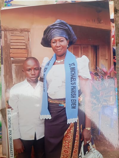 A medium shot photograph captures a woman and a boy standing in front of a building in Dutsen Alhaji, Nigeria. The woman, on the right, is wearing a black headwrap, a white ruffled blouse, and a dark skirt with a patterned trim. A blue sash draped around her neck reads "ST. MICHAEL'S PARISH EDEM." She has a watch on her left wrist and rings on her fingers, and is holding a silver purse. The boy, on the left, is dressed in a white collared shirt and dark pants. He has a serious expression. The background shows a weathered wooden door with louvers, part of a building wall, and some foliage. A handwritten sign above the woman's head reads "NO 99 EDEN 1". The bottom left of the image features a watermark that says "J.C DIGITAL PIX 08039464371".