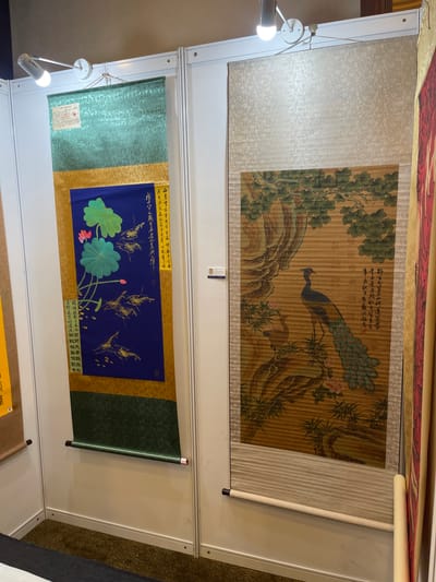 This media file captures an indoor exhibition display of traditional East Asian scrolls. Two large scrolls are prominently featured, hung vertically.
The upper scroll depicts a traditional painting of a peacock with its tail feathers fanned out, perched on a branch of a tree with lush green foliage and pink flowers. The painting is rendered on a textured, parchment-like background with vertical lines. A smaller, second peacock is visible in the lower right portion of the painting. Chinese calligraphy is present in the upper right corner of this scroll.
The lower scroll features a vibrant blue panel with a gold-patterned border. On the blue panel, there is a painting of lotus flowers and leaves, along with several depictions of shrimp in gold ink. Vertical lines of Chinese calligraphy are present in yellow borders on the top and bottom of the blue panel. To the left of this scroll, a textured green scroll with a red and white label is partially visible.
The scrolls are displayed against a white backdrop, with silver metal framing and lighting fixtures visible. The overall setting appears to be a well-lit exhibition hall, possibly a market or gallery, given the display method. The context suggests a cultural or art-related event, likely in Futian District, China, focusing on traditional scroll art. No people are visible in the frame.