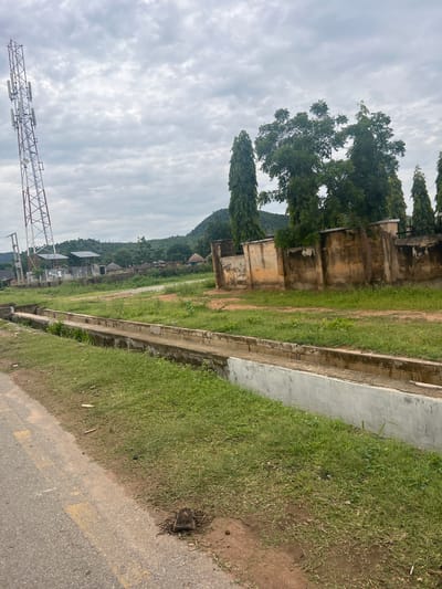 The image depicts an outdoor scene in Kona Garu, Nigeria, under a cloudy sky. The primary subjects are a concrete drainage channel, a grassy area, and a road with yellow markings. In the background, there are several structures including old, weathered buildings and tall, slender trees. Further in the distance, a lush green hillside with scattered structures can be seen. On the left side of the image, a large metal tower-like structure, possibly a communication tower or part of a construction crane, is visible against the sky. The overall setting appears to be a semi-urban or rural area. There is no clear activity or event taking place, and no people are visible. The time of day is indeterminate due to the overcast weather, but the lighting suggests it is daytime. The environment appears somewhat neglected, with peeling paint on the buildings and overgrown grass.
