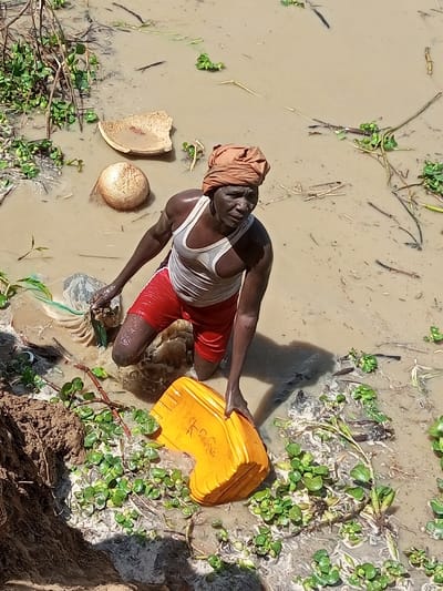 An adult male is observed kneeling in shallow, turbid, brownish-green water in Kokani North, Nigeria. He wears a light-colored sleeveless undershirt, red shorts, and a light brown cloth wrapped around his head. His gaze is directed towards the right side of the frame. He is actively pressing a bright yellow plastic container, possibly a jerrycan, into the water with both hands. His left hand is also positioned near some submerged debris. Floating in the water are several objects, including a fragmented, light-colored bowl, a spherical woven object, and numerous green aquatic plants (resembling water hyacinth) mixed with dried vegetation and other debris. A mound of dark brown earth is visible at the lower-left edge of the water, indicating the bank. The scene is illuminated by bright sunlight, casting shadows.