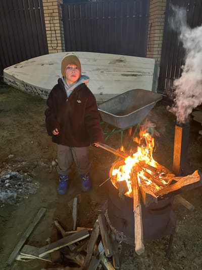 A person stands in a dirt-covered yard, next to a burning fire in a black drum. The individual wears a brown jacket, gray pants, a hat, and blue boots. Wood is stacked on the ground near the fire. Behind the person, a white boat rests on its side, with visible lettering in Russian. A wheelbarrow sits next to the boat. A fence and brick wall are visible in the background. Smoke rises from the fire. Patches of snow or ice are scattered on the ground. The location is слобода Старая, Russia.