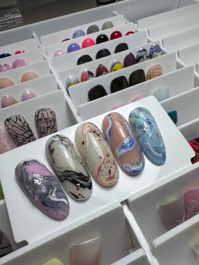The image displays an overhead view of organized white trays filled with artificial fingernails, each showcasing distinct designs. The fingernails are arranged in rows and columns within individual compartments. The designs include marble patterns in various color combinations such as pink and gray, black and white, and blues and greens, some with added glitter or foil flakes. Other designs feature intricate black line art on a pink background and a web-like pattern on a pinkish nail. In the upper portion of the image, there are rows of smaller, rounded objects, possibly polish caps or small nail art embellishments, in a variety of colors including blue, pink, red, and grey. The overall setting appears to be a professional nail art studio or a personal collection of nail supplies. The location of Vlasikha, Russia, is not depicted or suggested by any visual elements in the image.