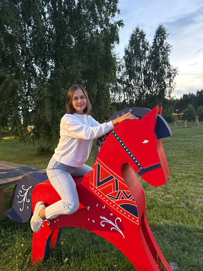 A young woman is posing for a photo, sitting on a large, red, wooden horse sculpture. The sculpture is painted with dark blue and white accents and decorative patterns. The woman is wearing a white sweatshirt and light blue jeans. She is smiling at the camera.

The background shows a grassy field with trees. The sky is cloudy, suggesting it is late afternoon or early evening. The lighting is soft, and there are some blurred lights in the distance, possibly from lamps or fairy lights. The overall atmosphere is relaxed and cheerful. The setting appears to be an outdoor park or recreational area. There is no visible text in the image.