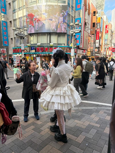 In Shibuya, Japan, on a busy street lined with bright neon signs and billboards, a young woman in a white Lolita-style dress with tiered ruffles and intricate white lace stockings stands facing away from the camera. She wears platform boots and holds a smartphone in her right hand, appearing to take a photo or video. To her left, an older woman in a black jacket and dark pants gestures animatedly with her hands and holds a bouquet of pink roses in a clear plastic wrapper. She is looking at the younger woman. In the background, other pedestrians navigate the crowded sidewalk. The street is paved with herringbone-patterned bricks, and tall buildings with numerous signs in Japanese script dominate the cityscape. One prominent billboard displays a colorful image of people in traditional costumes.