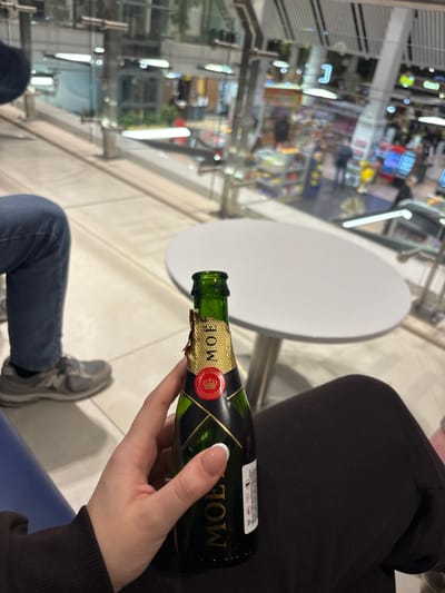 An indoor public facility, located in Bubuieci, Moldova, is depicted. The scene is viewed from a slightly elevated position within a modern, multi-level structure featuring glass partitions and metal framing. In the immediate foreground, a human right hand, with manicured fingernails, holds an opened miniature green glass bottle of Moët & Chandon champagne. The bottle's gold foil cap is partially torn. To the left, the lower body of another individual is visible, wearing blue jeans and grey New Balance athletic shoes, indicating a seated posture. A small, round, white table with a metallic pedestal base stands empty between the subjects. Beyond the glass partition in the background, a lower level of the facility is visible, featuring various retail establishments with brightly lit shelving and blurred human figures indicative of pedestrian traffic. An escalator is also discernible in the background. The lighting is artificial and bright throughout the facility.