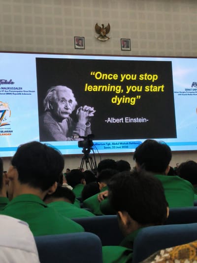 The image shows a screen displaying a quote by Albert Einstein: "Once you stop learning, you start dying." The quote is presented alongside a black and white image of Einstein. The screen also shows text in Indonesian, including the name "MALIKUSSALEH" and details about an academic event, stating it is the 57th Scientific Oration and Presentation in the framework of the National (BRIN) Republic of Indonesia. Below the quote, it reads "-Albert Einstein-". Further down the screen, a date "Senin, 22 Juni 2026" and location "Auditorium Tgk. Abdul Wahab Dahlawy" are visible. The scene takes place in an auditorium with an audience, mostly young people wearing green shirts, seated in blue chairs, facing the screen. The background wall has a grid pattern and features two framed pictures and an eagle emblem. A tripod with a camera is positioned in front of the screen, indicating a recording or presentation. The overall atmosphere appears to be formal and educational. The location is explicitly stated on the screen as Auditorium Tgk. Abdul Wahab Dahlawy, which is part of Malikussaleh University, located in Reuleut Timu, Indonesia.