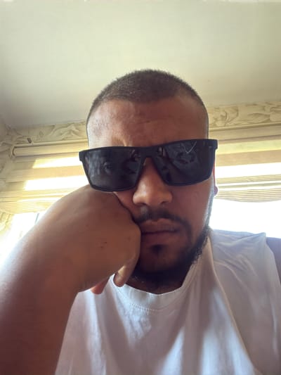A selfie taken from a low angle shows a man wearing sunglasses and a white tank top. He rests his head on his left hand, with his elbow on a surface. The man has short, dark hair and a beard. He is looking directly at the camera. In the background, blinds are visible, suggesting an indoor setting. The lighting indicates it is daytime. The image appears to be taken in Kalitinovo, Bulgaria.